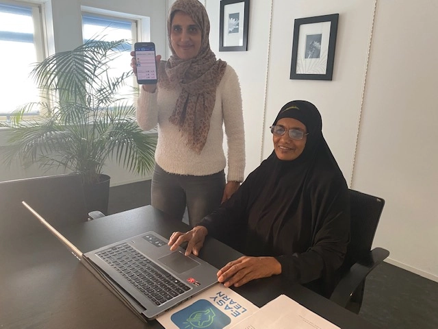 To indvandrekvinder. den ene står op og holder en mobil op den anden sidder foran en bærbar computer
